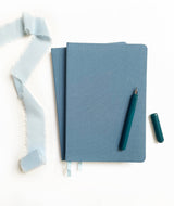 Two Linen Hardcover Journals, a teal fountain pen with cap, and blue fabric on a white background.