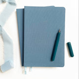 Two Linen Hardcover Journals, a teal fountain pen with cap, and blue fabric on a white background.