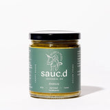 A jar of Zhough Dip & Spread, a spicy cilantro jalapeño sauce with a green label, sits on a white surface.