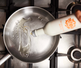 A hand pours Algae Cooking Oil from a white bottle into a stainless steel pan on a gas stove.