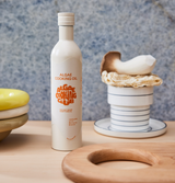 A bottle of Algae Cooking Oil sits on a wooden table beside stacked dishes and a mushroom on a plate.