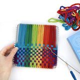 Hands weaving rainbow cotton loops on a 7 inch metal traditional potholder loom, with a pile of colorful loops nearby