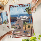 Two people assemble the 1000 Piece Santa Barbara Stern's Wharf Puzzle with drinks on a light-colored table.