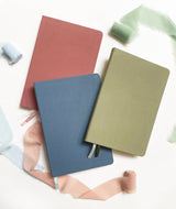 Three Linen Hardcover Journals in red, blue, and green, displayed on white with pastel fabric strips around them.