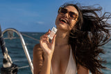 Woman in sunglasses smiles on a boat, holding Tinted Sunscreen Stick SPF 30, her windswept hair under the sun.