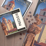 The 1000 Piece Santa Barbara Mission Puzzle is partially assembled next to its box and posters on a wooden table.