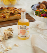 A bottle of Roasted Garlic Oil with Yuzu & Miso sits on a table near garlic, salad, bread, and a beige napkin.