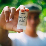 Hand holding a stick of sunscreen labeled 'SIN-MIN Shade' with a blurred background