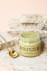 A jar of Circular Living Balancing Face Mask, made with matcha and French green clay, rests on textured stone slabs next to two gold measuring spoons. The mineral-rich mask powder inside is a pale green shade.