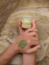 A person holds a Circular Living Balancing Face Mask jar in one hand, while the green clay mask is applied to the back of their other hand. The background features textured stone.