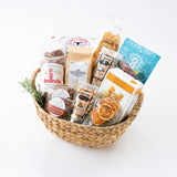 The California Farm Snacks Gift Basket features a woven basket filled with almonds, dried fruit, pasta, crackers, and cookies. All items are arranged on a white background with sprigs of rosemary at the front.