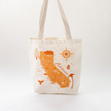 The California Map Tote is a cream recycled canvas bag with beige handles, featuring an orange map design with icons like a bear and whale tail—a charming gift or wedding bag perfect for any California enthusiast.