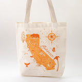 The California Map Tote is a cream-colored bag with orange illustrations of the state map, landmarks, animals, sun, trees, and “California” in cursive on recycled canvas—ideal as a unique wedding gift bag.