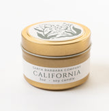 A gold tin labeled “Santa Barbara Company California Travel Candle” (3oz soy wax) features a black and white floral illustration on the lid. The travel-friendly candle rests on a white surface.