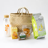 The California Welcome Gift Basket features a woven straw bag with a ribbon handle, plus a floral white water bottle, yellow gift box, dried apricots, almonds, and sea salt & olive oil chips for the perfect California treat.