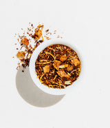 A white bowl of Caramelized Pear Loose Leaf Tea with dried fruit sits on a white surface, some pieces spilled beside.