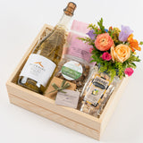 The Cheers Celebration Gift Box includes white wine, gourmet treats, chocolates, and a vibrant orange and purple floral arrangement, all beautifully arranged in a wooden box with shredded paper for an elegant presentation.