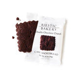 A square, scalloped-edge Chocolate Cacao Nib Shortbread Cookie rests partly outside its white Rustic Bakery wrapper. Cookie crumbs and cacao nibs are scattered nearby.