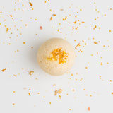 The Citrus Sunshine Bath Bomb, round and beige with an orange powder center and citrus oils, rests on a white surface amid small orange and beige crumbs.
