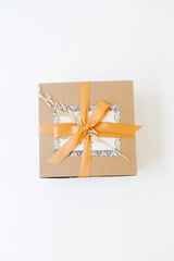 The Classic California Welcome Gift, a brown box with an orange ribbon, dried wheat accent, and a patterned card, sits on a white background—ideal for self-care or sharing California snacks.