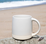 The Custom Logo Coronado Ceramic Mug 12 oz rests on a sandy beach rock, with blurred ocean waves and shoreline in the background—a peaceful scene ideal for displaying your personalized mug.
