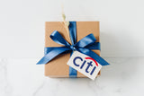 A brown gift box topped with a blue satin ribbon and dried grass showcases a Custom Logo Gift Tag on a white marble surface.