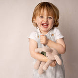 Smiling toddler in striped overalls holds a Fair Trade Handmade Bunny in Suspenders
