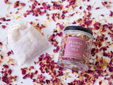 The Floral Bath Tea comes in a reusable glass jar with aromatic dried organic florals. It includes a muslin bag for an inviting, relaxing bath soak. The product is displayed on a white surface with scattered petals.