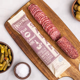 A wooden board with sliced Foustman's Pork Cabernet Salami, its packaging visible, set beside dishes of olives, pickles, and a small bowl of salt on a white surface.