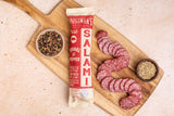 A wooden cutting board with Foustman's Pork Fennel & Pepper Salami, several slices of salami, a small bowl of mixed peppercorns, and a small bowl of fennel seeds on a light countertop.