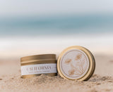 A Golden State Sunshine Gift Tote rests on beach sand by the ocean, its gold tin adorned with floral art. The softly blurred scene evokes a relaxed California vibe—making it a perfect local gift.