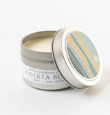The Goleta Bluffs Travel Candle is a 3oz soy wax candle in a small silver tin. Its lid, decorated with abstract colorful waves and a sun, is partially off to reveal white soy wax. The label notes Santa Barbara Co.