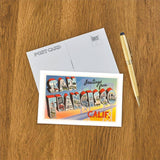 A Greetings from San Francisco Postcard rests by a pen on a wooden surface.