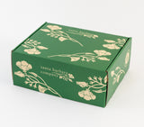 The Happy Place Handlebar Coffee Gift Box is a green cardboard box with cream floral designs and "Santa Barbara Company" on the lid and sides—perfect for gifting items like an insulated camp mug or Handlebar Coffee treats, shown against a plain white background.