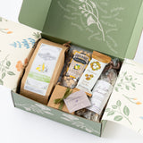 The Happy SB Treats Gift Box with Custom Logo features a green floral box filled with sea salt & olive oil crackers, organic pistachios, Santa Barbara snacks, a small wrapped package, and cookies all beautifully arranged.