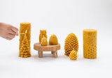 A hand lights a Hive Beeswax Candle shaped like a Christmas tree, surrounded by other natural candles—carved beeswax owls, pinecones, a floral pillar, and skep candles—displayed on a white background.
