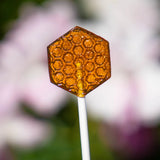 Honeycomb Lollipops are translucent amber hexagons with a raised honeycomb pattern, made from organic wildflower honey on white sticks, shown against a soft pink and white blurred background.