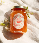 A jar of Ruby Valencia Orange and Apricot Marmalade with a label showing the brand Marmalade Grove placed on a linen surface, accompanied by apricot flowers.