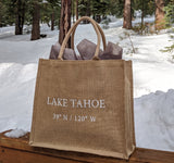 The Lake Tahoe Coordinates Jute Tote, featuring "LAKE TAHOE 39°N / 120°W" in natural jute, sits on a wooden surface amid snowy pines—a stylish and eco-friendly bag perfect for Lake Tahoe enthusiasts.