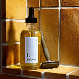 A bottle of Lavender Body & Bath Oil rests beside a metal safety razor on a sunlit tiled surface, with warm yellow-brown tiles in the background.
