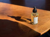 A small glass dropper bottle of Lavender Body & Bath Oil rests on a wooden table, casting a long shadow in the warm sunlight.