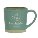 The Los Angeles Shells Etched Ceramic Mug is a branded 12 oz green ceramic cup with a beige base, featuring etched sea shells and "Los Angeles CALIFORNIA" in white on the front.