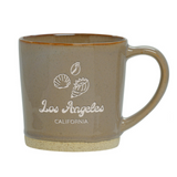 The Los Angeles Shells Etched Ceramic Mug is a beige 12 oz mug with a handle, featuring white seashell illustrations and "Los Angeles CALIFORNIA" on the front—ideal for enjoying your favorite drink in style.