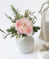 The Love Bud Vase, a small white vase, holds a pink ranunculus stem with green leaves and delicate white buds, with part of a beige fabric bag seen in the background on a white surface.