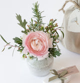 The Love Bud Vase holds a pink ranunculus stem, rosemary, and greenery. Beside it are a beige gift bag with a tag and a small box tied with green ribbon, all arranged in a bright, minimal setting.