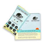 An Ocean Beach Omega San Diego Snack Bar sits atop its 8-pack box. The packaging features a beach theme, highlights plant-based, omega, and non-GMO ingredients, and includes a message supporting the community.