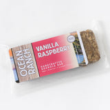 A packaged Ocean Ranch Handcrafted Granola Bar labeled Vanilla Raspberry, highlighting 11g vegan protein, wheat-free, and locally crafted.