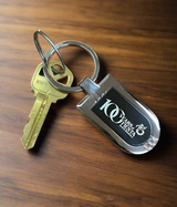 A brass house key on a keyring with the Official Fiesta Centennial Keychain, which reads "100 Years Fiesta 1923-2023," rests on a wooden surface, commemorating the 100th anniversary.