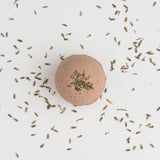 The Ojai Lavender Bath Bomb is a round brown bath bomb topped with dried lavender, infused with soothing jojoba oil, resting on a white surface surrounded by scattered lavender buds.