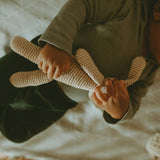 Baby lying down holding an Organic Cotton Bunny rattle, shown from the shoulders down on a patterned blanket.